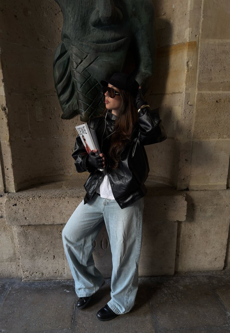 Frau mit Sonnenbrille, schwarzer Lederjacke und Handschuhen, hält Zeitschriften und steht an einer Steinmauer unter einer großen geschnitzten Gesichtsskulptur.