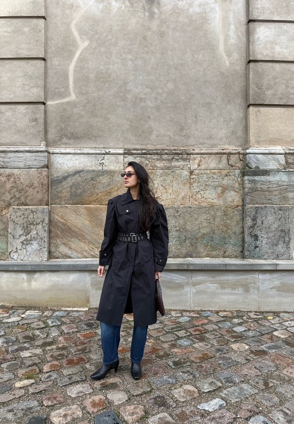Una mujer con un abrigo negro con cinturón, vaqueros y gafas de sol está de pie en una calle empedrada junto a un gran edificio de piedra.