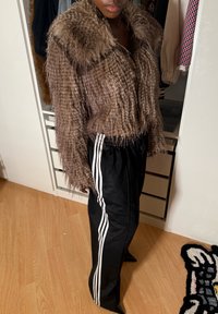 Personne portant une veste brune en fausse fourrure et un pantalon de survêtement noir avec des bandes blanches, debout dans une pièce avec un sol en bois et un placard ouvert.