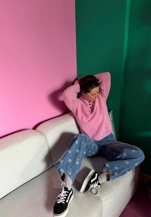 Persona sentada en un sofá gris claro, vistiendo un suéter rosa, jeans azules con lunares, zapatillas negras, en una habitación con paredes rosas y verdes.