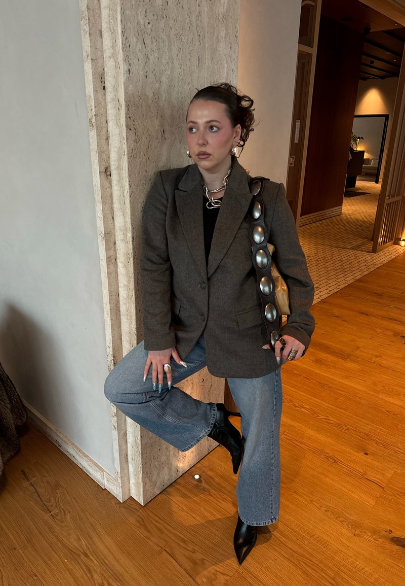 Junge Person trägt ein graues Sakko, blaue Jeans und schwarze Stiefel, lehnt sich an eine beige Steinsäule in einem modernen Innenraum.