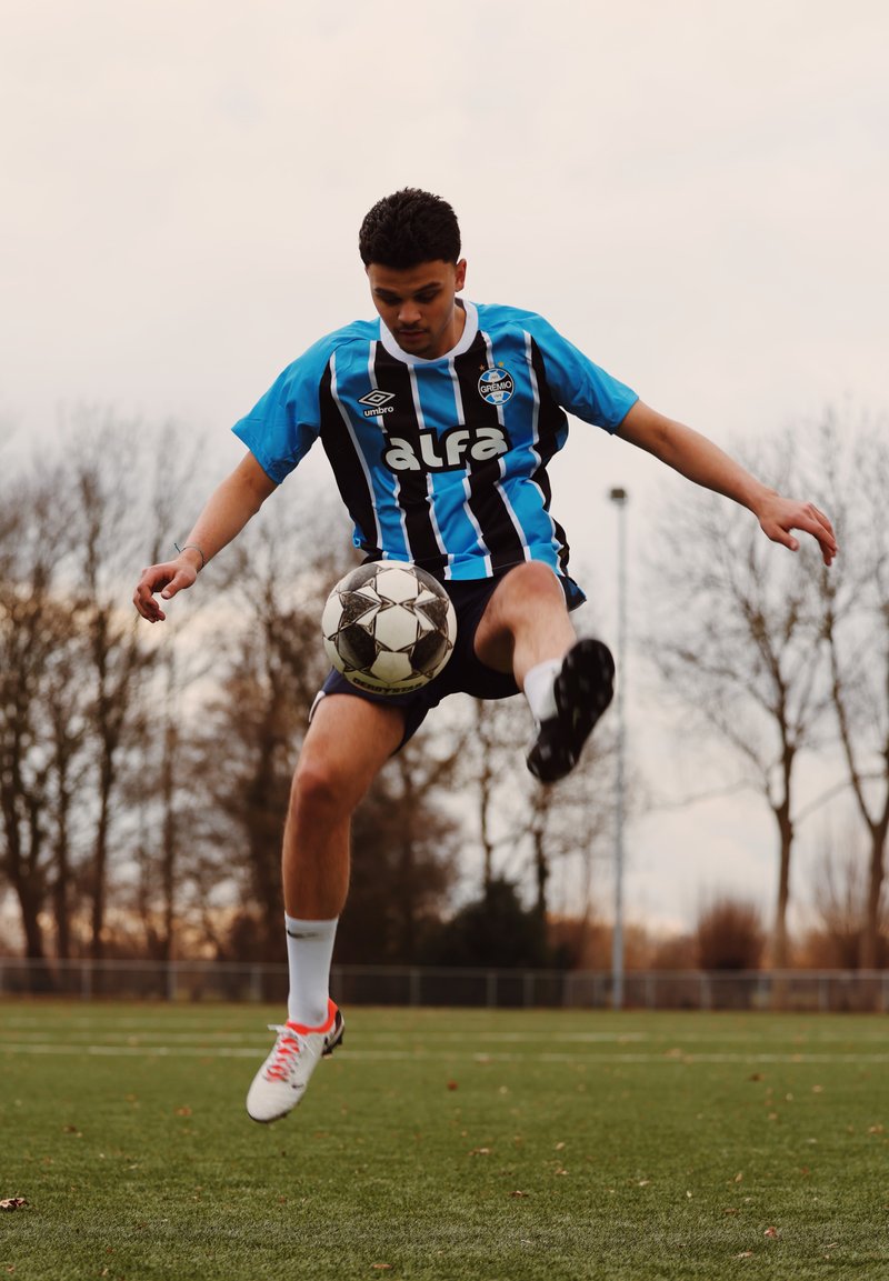 Jeune joueur de football masculin en maillot à rayures bleues et noires contrôle le ballon en l'air sur un terrain extérieur avec des arbres nus en arrière-plan.