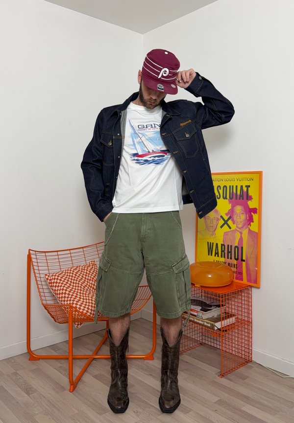 Hombre ajustándose una gorra burdeos, con chaqueta vaquera, camisa blanca con estampado de velero, pantalones cortos cargo y botas vaqueras en una esquina con silla naranja y póster artístico.