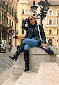 Cappotto trench in pelle nera, occhiali da sole neri, stivali neri fino al ginocchio, jeans blu e una borsa a tracolla bianca, seduto su un gradino di pietra in un contesto urbano.