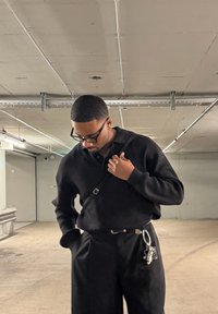 Čierny pletený sveter a čierne nohavice v parkovacej garáži. Sveter má golier a je viditeľná kabelka cez rameno s kovovými detailmi.