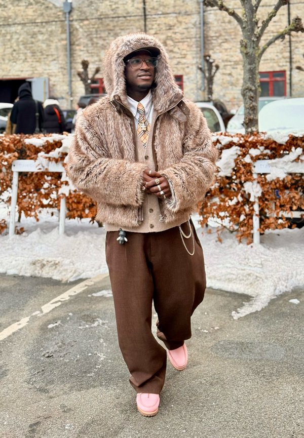 Moški z očali, bež jakno s kapuco iz umetnega krzna, s kravato z vzorcem, rjavimi širokimi hlačami in roza čevlji, stoji na zasneženi tlakovanih površini blizu žive meje.