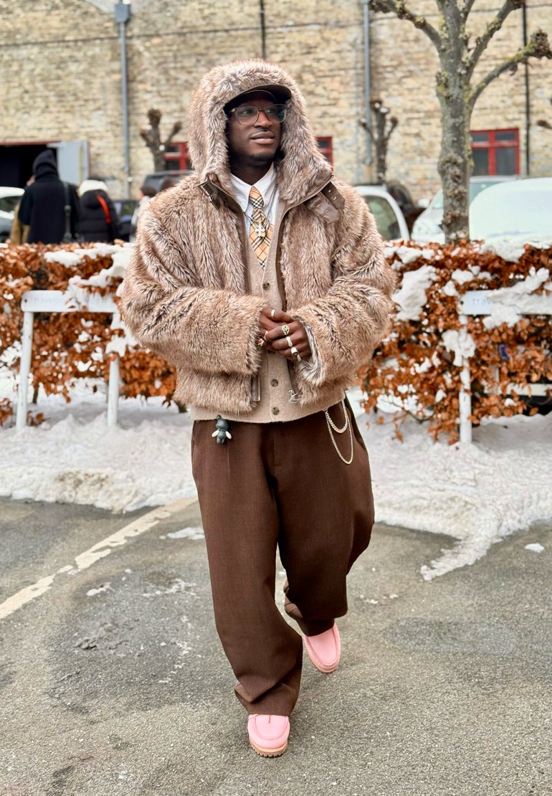 Homme portant des lunettes, une veste beige à capuche en fourrure, une cravate à motifs, un pantalon large marron et des chaussures roses, debout sur un trottoir enneigé près d'une haie.