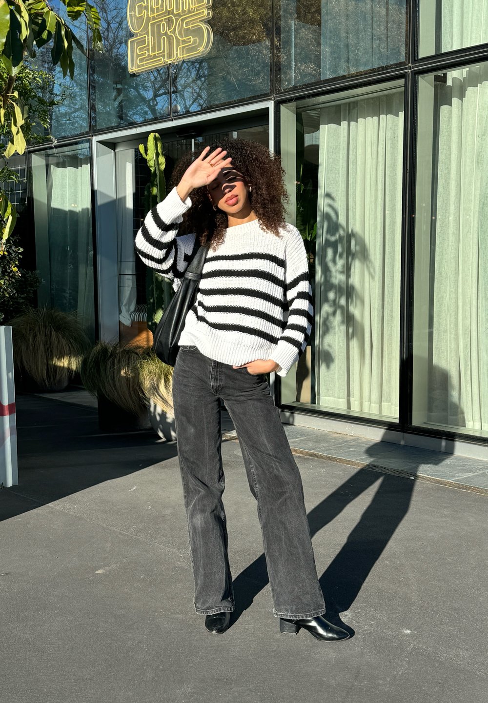 Randig svart och vit tröja, avslappnad passform; svarta vida jeans; svarta ankelstövlar; står utomhus i solljus med en byggnad i bakgrunden.