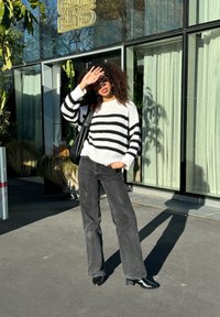 Randig svart och vit tröja, avslappnad passform; svarta vida jeans; svarta ankelstövlar; står utomhus i solljus med en byggnad i bakgrunden.