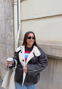 Femme portant une veste en peau lainée noire, un pull rayé, des lunettes de soleil, tenant une tasse de café et un sac fourre-tout crème, debout près d'un mur en béton.