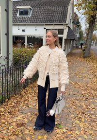 Béžová krémová bunda s texturou a chlupatým povrchem, béžový svetr na knoflíky, tmavě rozšířené džíny a světle šedá kabelka. Na pozadí podzimní listí.