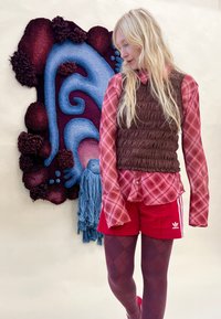 Blonde nő piros kockás ingben, barna mellényben, piros Adidas rövidnadrágban és mintás harisnyában áll egy textúrázott falművészet mellett, amely kék és bordó örvényeket ábrázol.