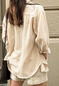 Striped beige button-up shirt with rolled sleeves, lightweight fabric, and a curved hem, paired with light-colored linen shorts.
