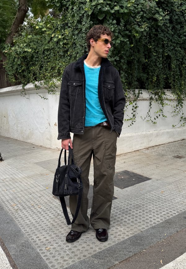 Joven con gafas de sol, chaqueta negra, camisa azul verdosa y pantalones oliva, sosteniendo una bolsa negra, de pie en una acera con vegetación detrás.