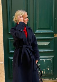 Une femme blonde ajuste ses cheveux d'une main, portant un manteau noir par-dessus une chemise rouge, debout devant une grande porte en bois verte.