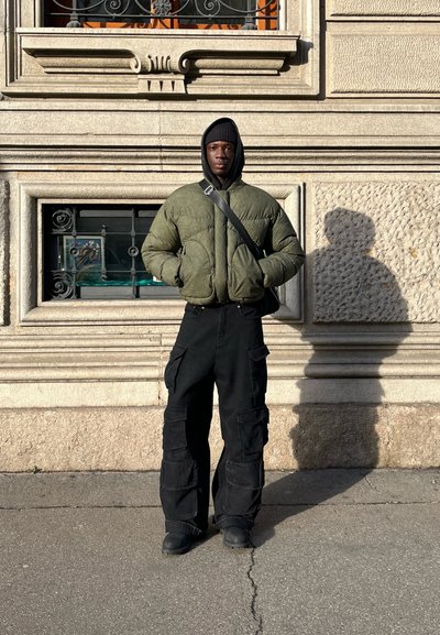 Chaqueta de abrigo verde con cierre de cremallera y bolsillos, combinada con pantalones cargo negros de corte holgado. Sombra proyectada en el pavimento.