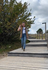 Chaleco sin mangas beige sobre camiseta blanca, jeans azules y zapatillas negras. Paseo de madera con barandillas, rodeado de vegetación y cielo nublado.