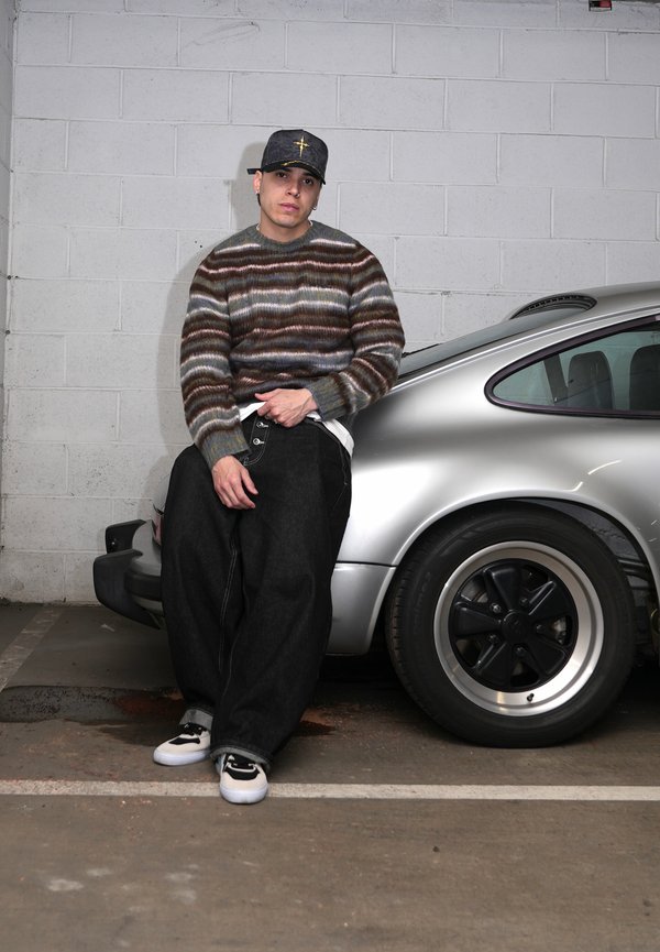 Ung mann i stripet genser, svart caps og vide jeans som lener seg mot en sølvfarget veteranbil i et parkeringshus.