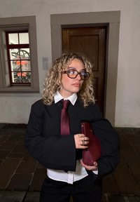 Czarna, oversizeowa marynarka z dużymi rękawami, biała koszula z kołnierzykiem, bordowy krawat i kopertówka. Kręcone blond włosy i przezroczyste okulary. Tło z kamiennej ściany.