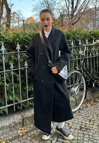 Lange zwarte double-breasted jas met grote knopen, gedragen over een wit overhemd. Gecombineerd met zwarte sneakers en witte sokken met structuur.