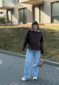 Jonge vrouw in een zwarte pet, donker langarmshirt, lichtblauwe jeans en witte sneakers staat op het trottoir met een bruine handtas.