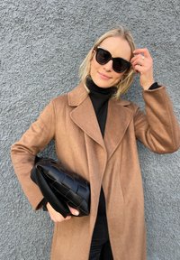 Brauner Wollmantel mit großen Revers, schwarzer Rollkragenpullover und übergroßer schwarzer geflochtener Clutch, vor einer strukturierten grauen Wand.