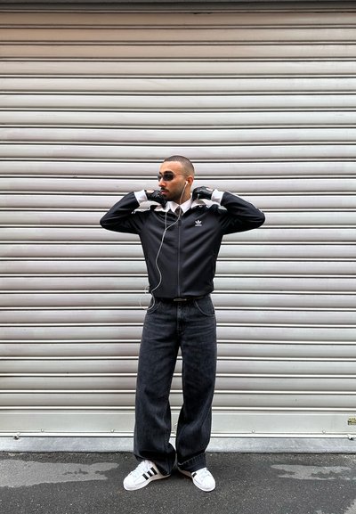 Hombre con chaqueta deportiva negra de Adidas, vaqueros negros, zapatillas blancas de Adidas, gafas de sol y guantes, ajustándose el cuello frente a una persiana metálica cerrada.
