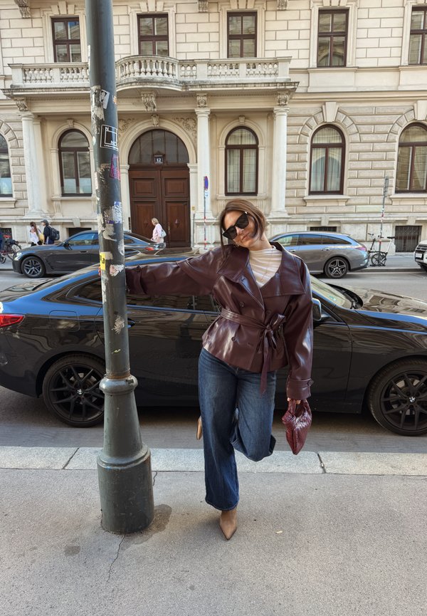Mujer con chaqueta oscura, vaqueros y gafas de sol se apoya en una farola con un pie levantado, de pie en una acera de la ciudad frente a coches aparcados.