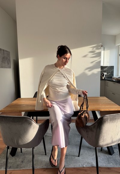 Mujer con suéter claro y falda lila sentada en una mesa de comedor de madera, sujetando un bolso marrón, en una cocina moderna iluminada por el sol.