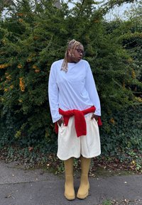 Fehér hosszú ujjú ing, vörös pulóver a deréknál megkötve, fehér rövidnadrág és sárga gumicsizma. A háttérben zöld lombok és narancssárga bogyók láthatók.