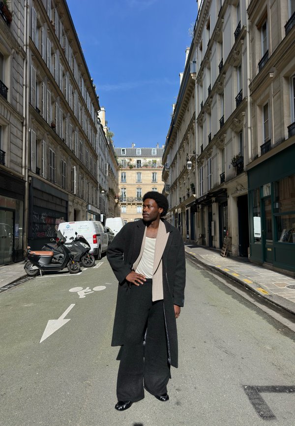 Mann iført svart kåpe og vide bukser står i en smal gate omgitt av fleretasjes parisiske bygninger under en klar blå himmel.