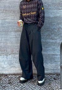 Μαύροι πλισέ παντελόνια με χαλαρή εφαρμογή, συνδυασμένα με πουκάμισο μακρυμάνικο σε σκούρο σχέδιο αργάιλι με χρυσές λεπτομέρειες και λογότυπο.