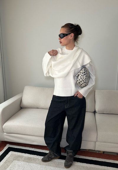 Mujer con gafas de sol negras, vestida con suéter blanco y pantalones negros holgados, está junto a un sofá beige sosteniendo un bolso de mano con manchas con una bufanda blanca.