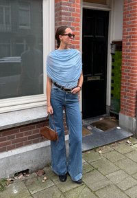 Jasnoniebieska bluzka w drapowania z drobnymi akcentami, zestawiona z spodniami dżinsowymi o wysokim stanie i rozszerzanymi nogawkami, brązową skórzaną torebką oraz czarnymi botkami na obcasie.
