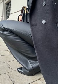 Schwarze Lederhosen mit glatter Textur, kombiniert mit einem langen schwarzen Mantel mit Knöpfen und einer kleinen schwarzen Handtasche mit goldenen Akzenten.