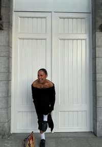 Zwarte off-the-shoulder trui met ribbelstructuur, gecombineerd met wijde broek. Witte sokken en donkere instappers. Geweven tas op de grond.