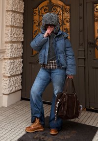 Džínová péřová bunda, šedý svetr, károvaná košile, modré džíny, hnědé boty a tmavě hnědá kabelka s kovovými doplňky, stojící u stylových dveří.