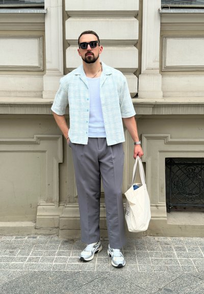 Camisa corta de manga azul claro con textura sobre una camiseta blanca, pantalones grises y zapatillas blancas. Sosteniendo una bolsa tote beige.