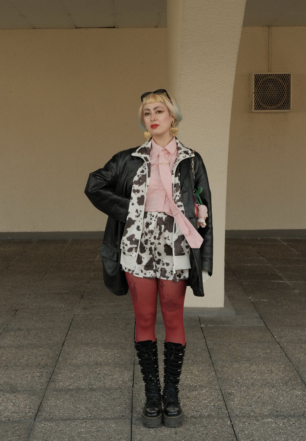 Persoon met kort blond haar en een zonnebril draagt een zwart leren jack over een koeienprintoutfit, rode panty’s, zwarte laarzen en hartvormige oorbellen.