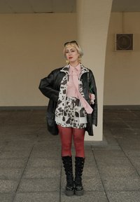 Persoon met kort blond haar en een zonnebril draagt een zwart leren jack over een koeienprintoutfit, rode panty’s, zwarte laarzen en hartvormige oorbellen.