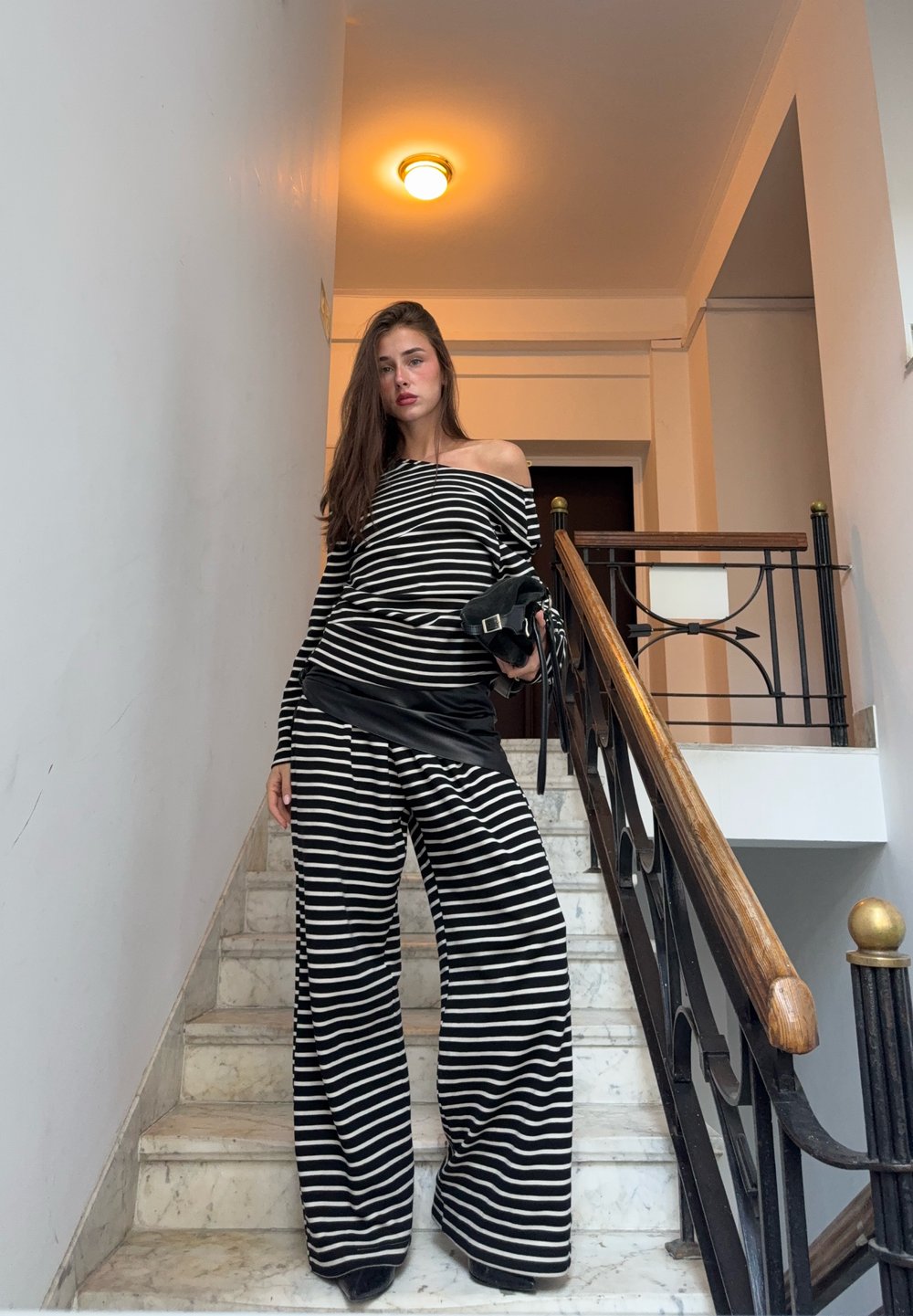 Haut rayé noir et blanc, épaules dénudées, et pantalon à jambes larges ; le mannequin tient un petit sac noir, debout sur un escalier en marbre.