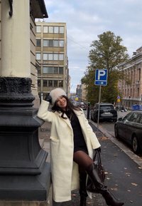 Donna con cappotto bianco e cappello di pelliccia appoggiata a una ringhiera di pietra su un marciapiede cittadino, con automobili e edifici sullo sfondo.