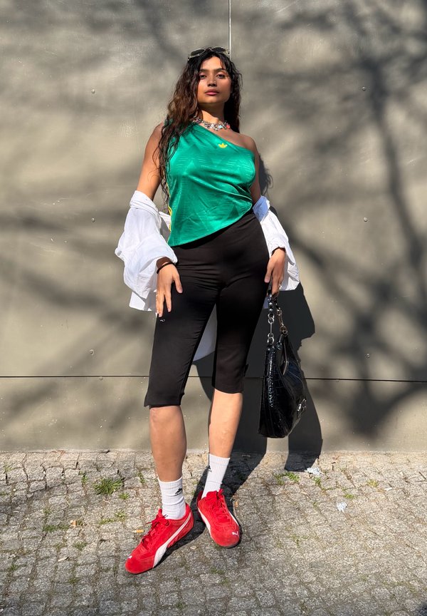 Vrouw staat tegen een muur, draagt een groene top met één schouder, zwarte capri-broek, rode sneakers, witte sokken en houdt een zwarte handtas vast.