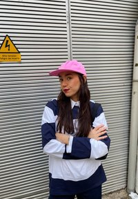 Ριγέ λευκό-μπλε πουκάμισο με μακριά μανίκια, ροζ καπέλο, στέκεται με τα χέρια σταυρωμένα μπροστά από ένα ασημί ρολό.