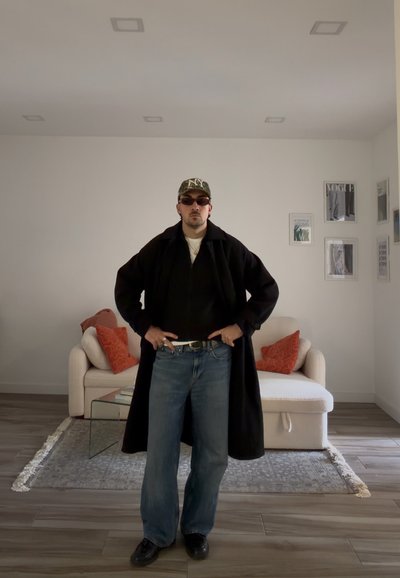 Hombre con abrigo negro, vaqueros azules, gorra estampada y gafas de sol de pie en una sala de estar con un sofá blanco y cojines rojos.