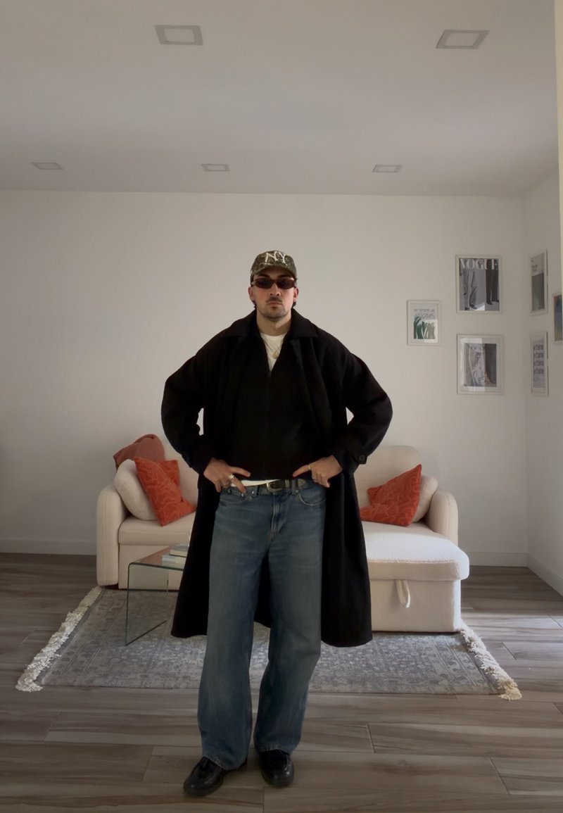 Mand iført en sort frakke, blå jeans, mønstret kasket og solbriller, stående i en stue med en hvid sofa og røde puder.