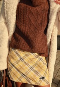 Pruuni palmikultuuriga kampsun beežist fliisjope all; kollase ruudulise mustri ja mustade ning valgete detailidega minisärk, millel on silt.