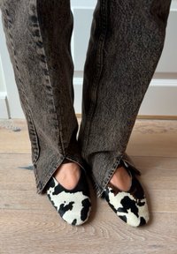 Zapatos tipo slip-on con estampado de vaca en blanco y negro, con un acabado texturizado, combinados con vaqueros acampanados grises que cuentan con aberturas laterales.