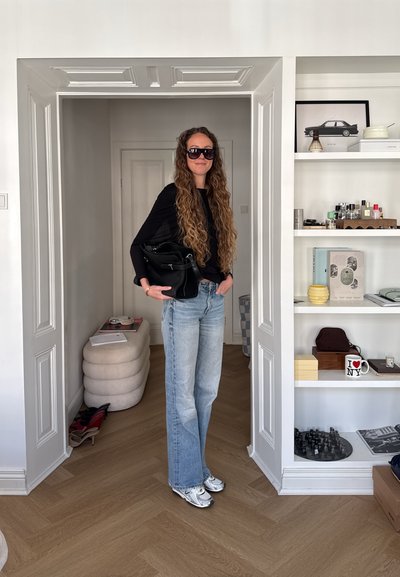 Mujer con pelo largo y rizado que lleva gafas de sol, top negro, vaqueros azules y zapatillas plateadas, de pie en una puerta sosteniendo un bolso negro.