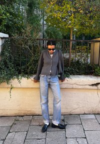 Γκρι καπιτονέ πουλόβερ με φουσκωτά μανίκια, λευκή μπλούζα, ανοιχτό μπλε φαρδιά τζιν και μαύρες εσπαντρίγιες. Πράσινη φυλλωσιά στο φόντο.